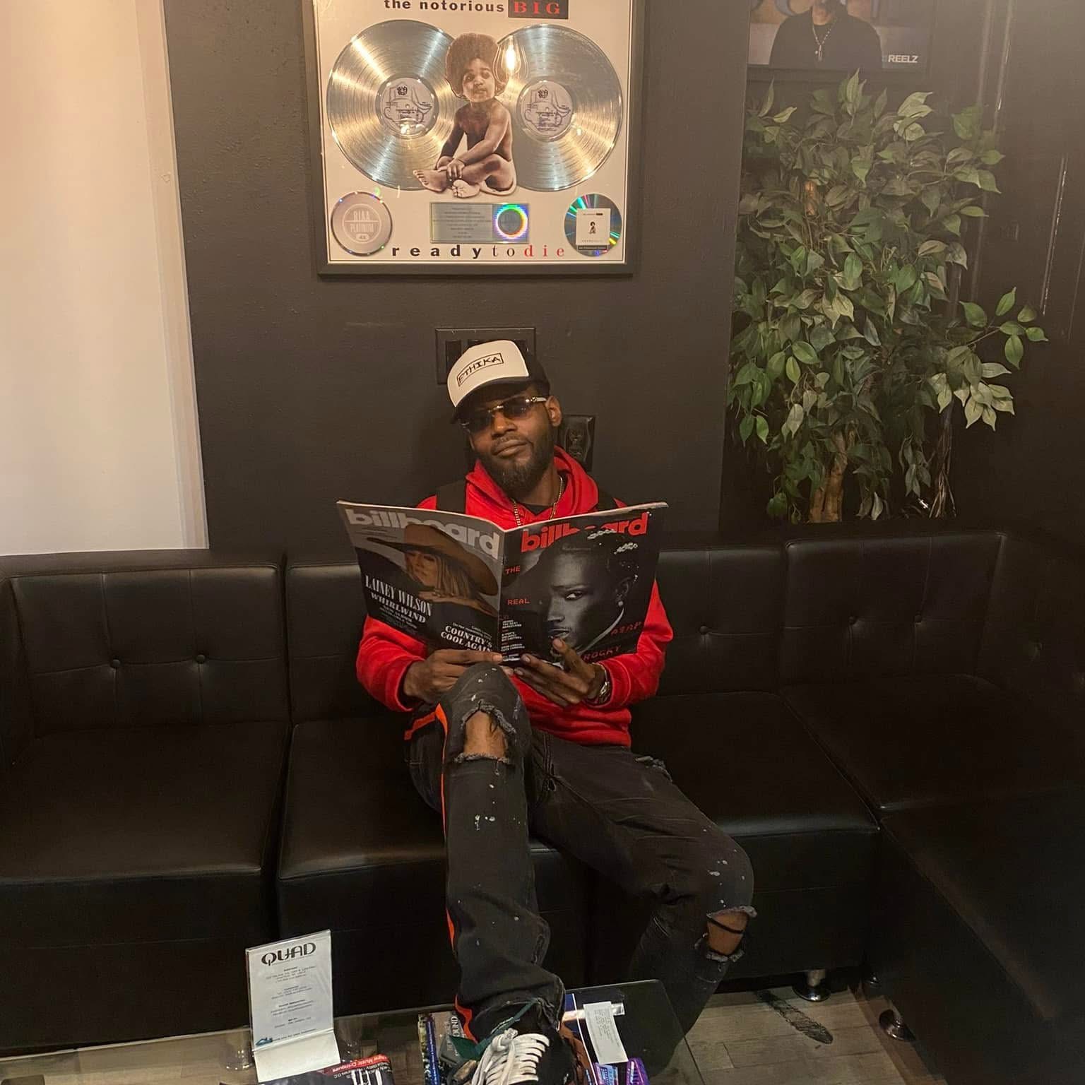 Flyboi Rich seated in a red hoodie holding a magazine.